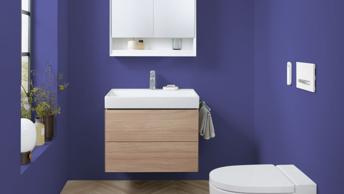 Pantone의 2022 올해의 색상인 "Very Peri"로 페인팅된 욕실에 있는 Geberit의 세라믹 제품과 욕실 가구가 있는 욕실.